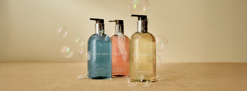 Drei Flüssigseifen von Molton Brown in transparenten Spenderflaschen stehen auf einer beigefarbenen Oberfläche. Die Farben der Seifen sind Blau, Rosa und Hellgelb. Um die Flaschen herum schweben zarte Seifenblasen, auf den Flaschen ist teils Schaum zu sehen. Die Szene wirkt sauber, frisch und hochwertig.