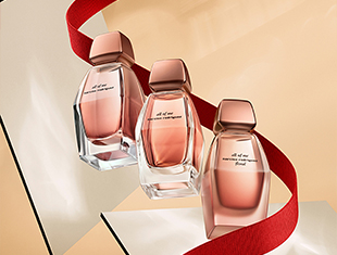 Drei ovale Parfumflakons der All of Me-Kollektion in Rosé- und Kupfertönen auf hellen Flächen arrangiert. Ein rotes Band schlängelt sich durch das Motiv und betont das festliche Design.