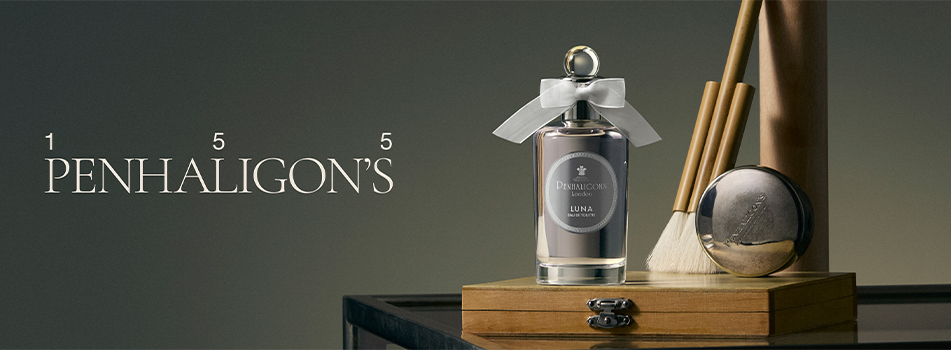 Penhaligon’s Parfumflakon 'Luna' steht auf einer Holzbox, umgeben von feinen Kosmetikpinseln und einer Puderdose. Der Flakon ist elegant mit einer weißen Schleife verziert. Links im Bild steht in stilvoller Schrift: 'Penhaligon’s 155'.
